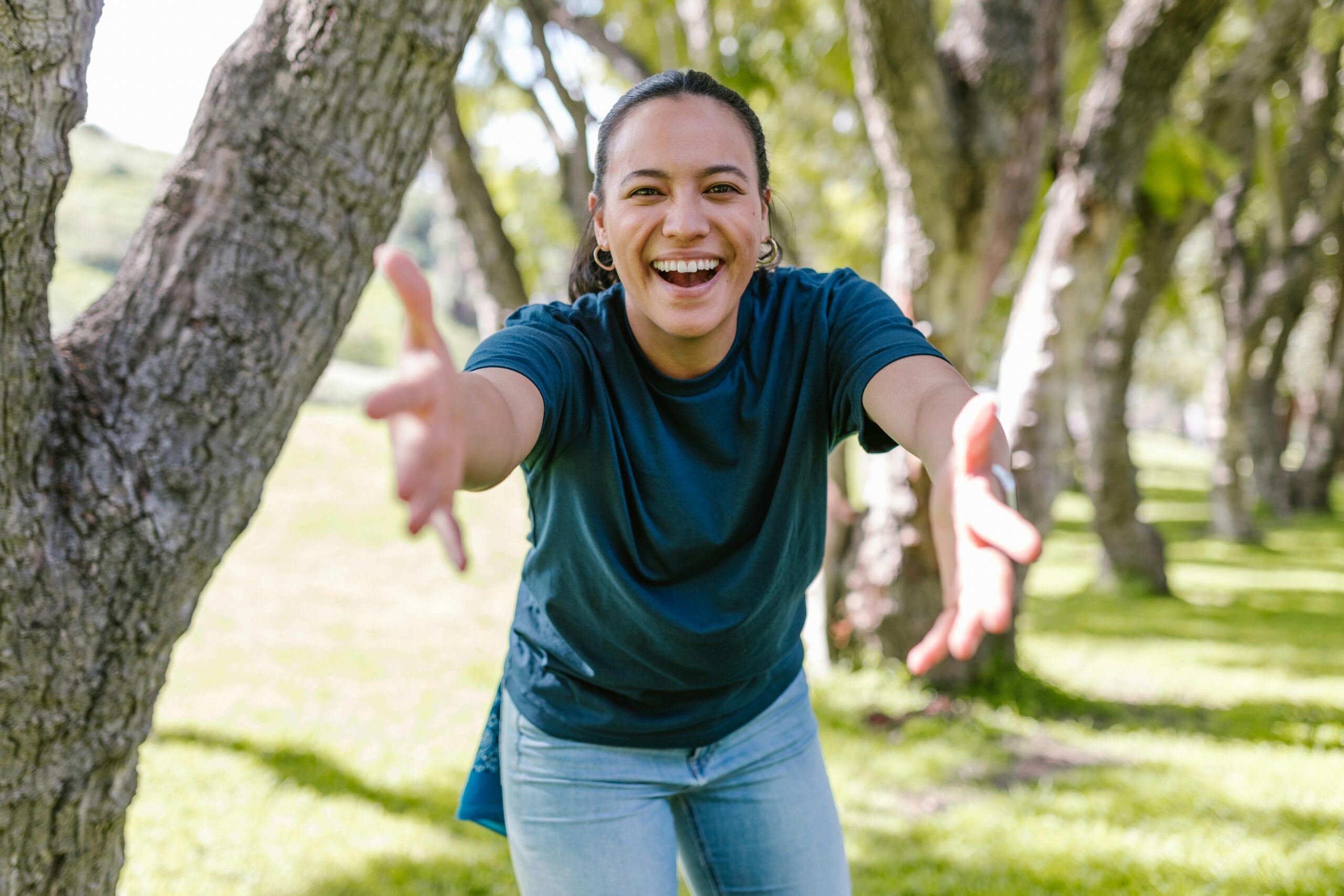 Smiling woman reaching out with open arms in a sunny, green park setting.