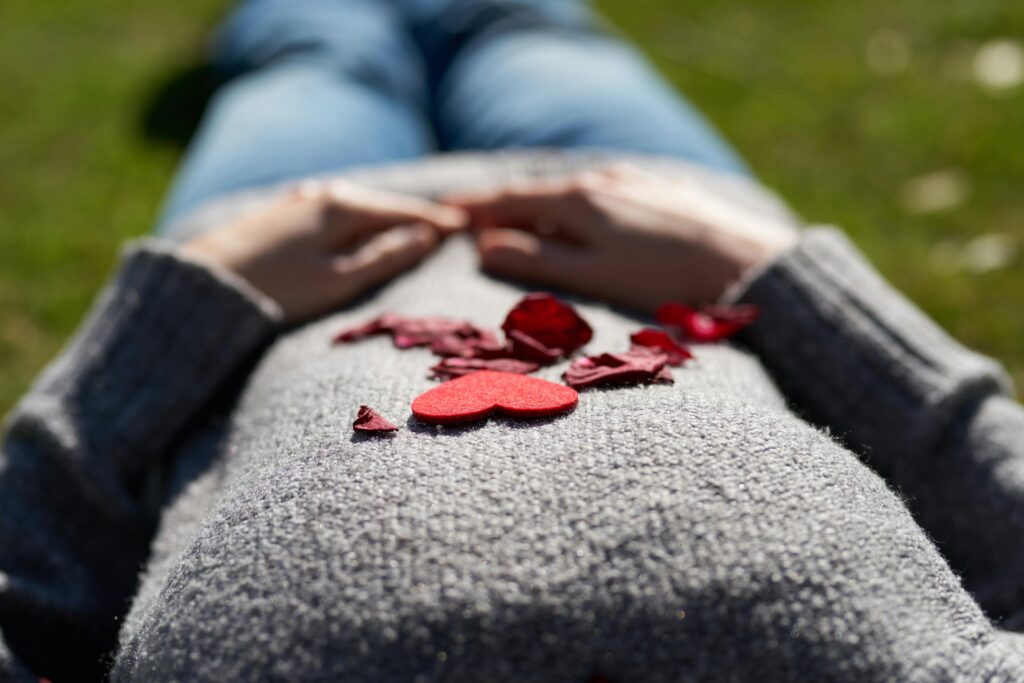 Pregnant woman lying on grass with heart and petals on belly, symbolizing love.